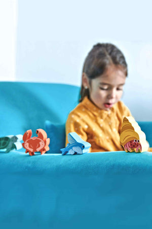 A child plays with colorful wooden coastal creature toys, including a crab and a fish, on a bright turquoise surface.