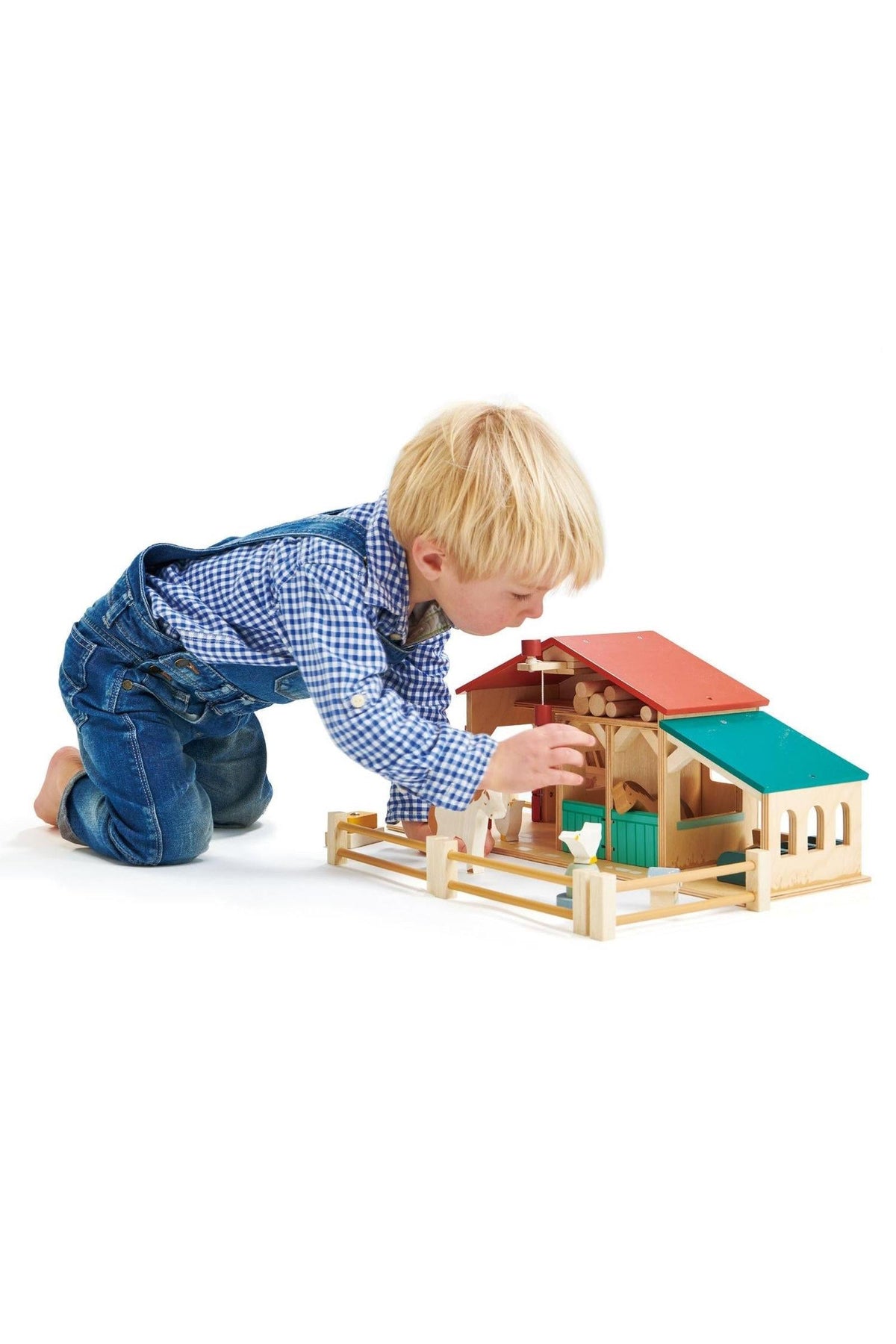 A young child kneels beside a wooden farm toy, interacting with its colorful barn and stables, promoting imaginative play.