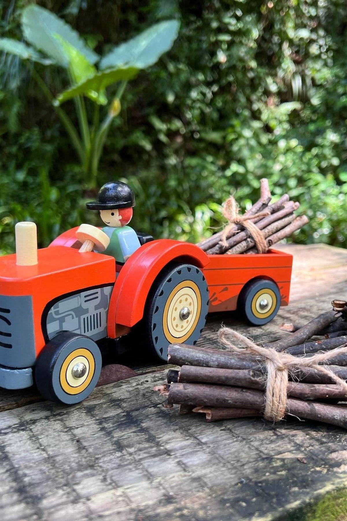 A vibrant red wooden tractor with a removable trailer, accompanied by bundled twigs, set against a lush green background.