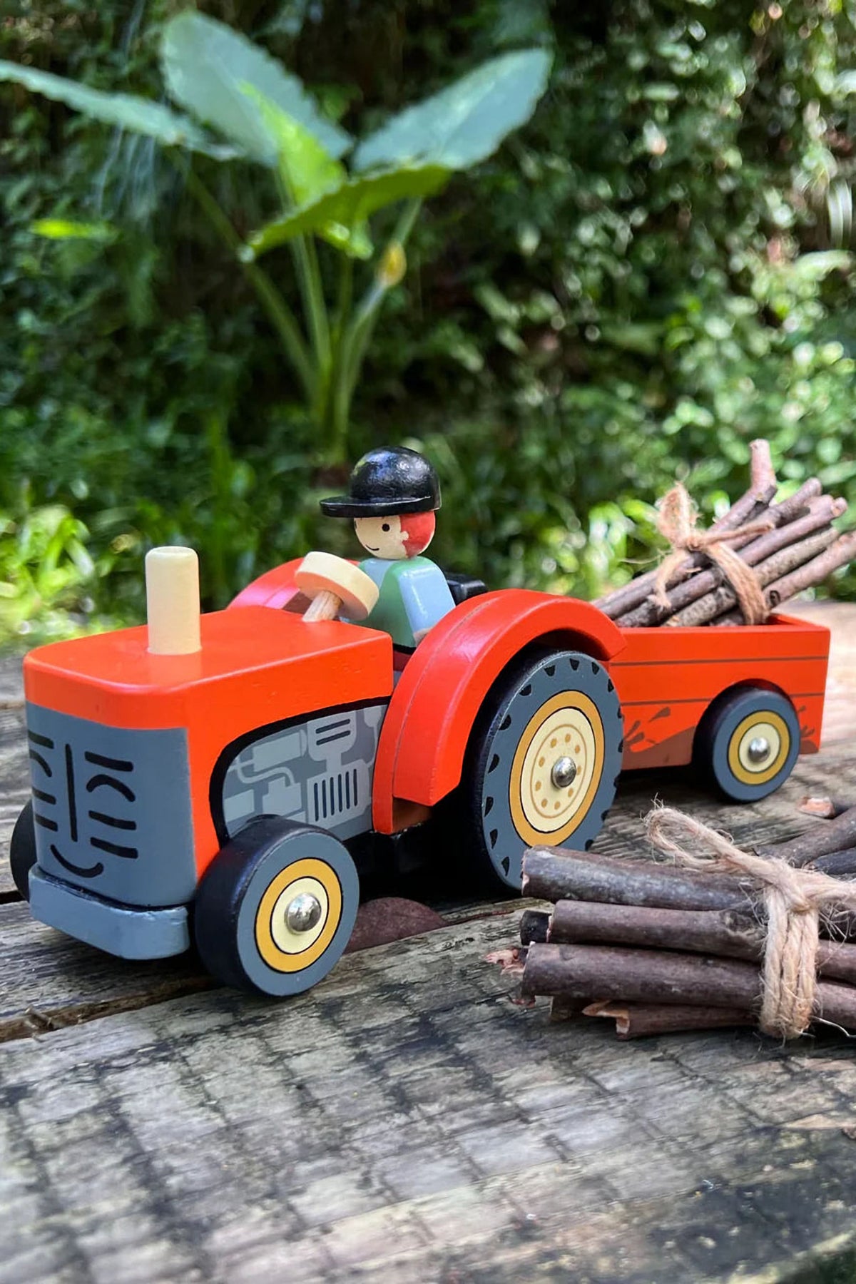 A vibrant red wooden tractor with a removable trailer carries bundled sticks, featuring a playful driver. This eco-friendly toy inspires imaginative play for young children in outdoor settings.