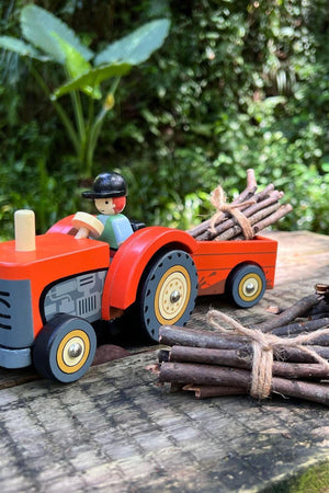 A vibrant red wooden tractor with a removable trailer, accompanied by bundled twigs, set against a lush green background.