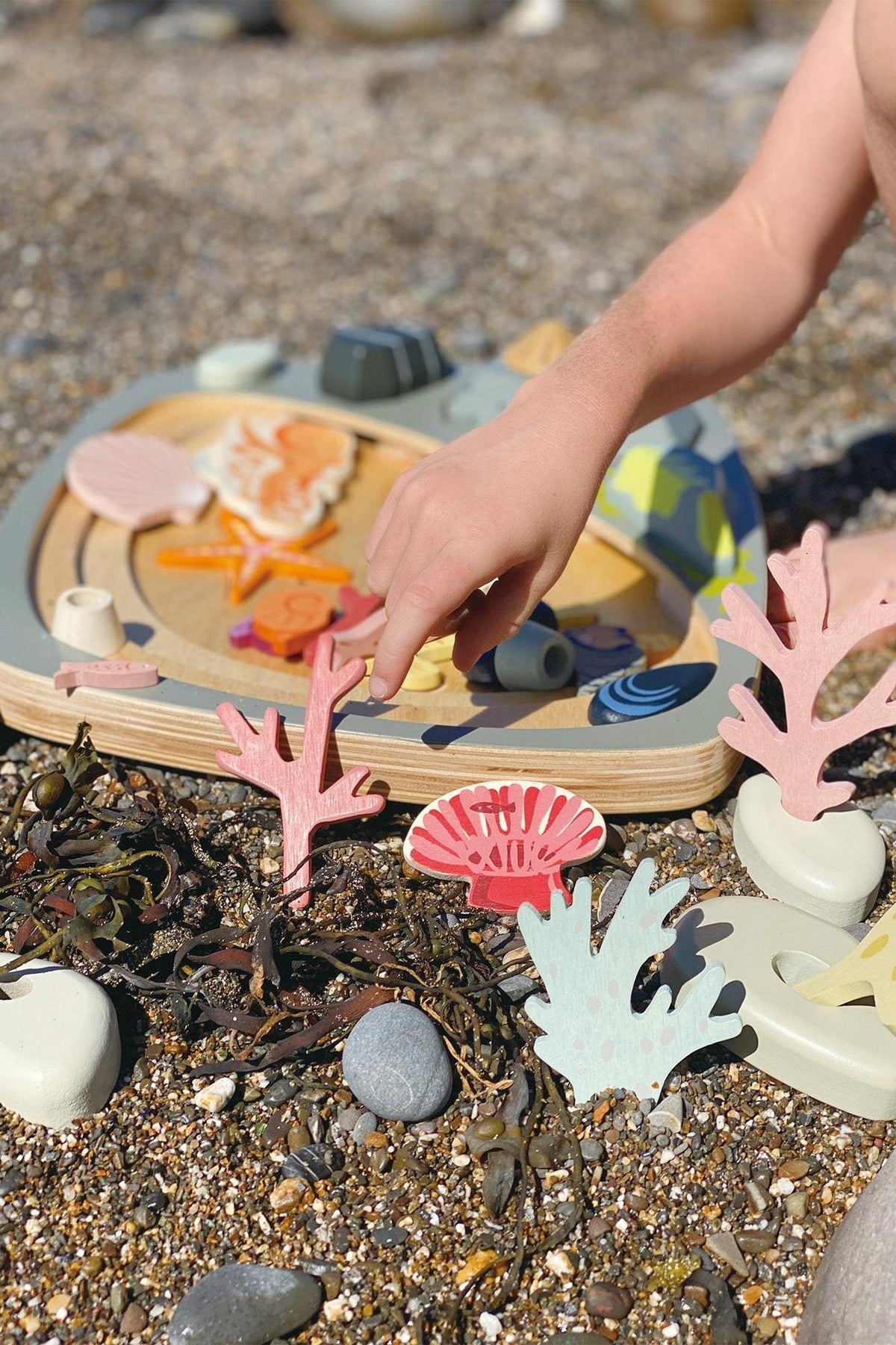 A child’s hand interacts with a colorful wooden shape-sorting toy surrounded by pebbles and seaweed. The toy features various marine shapes in pink, blue, and orange hues, highlighting eco-friendly materials.