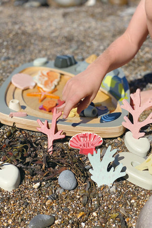 A child’s hand interacts with a colorful wooden shape-sorting toy surrounded by pebbles and seaweed. The toy features various marine shapes in pink, blue, and orange hues, highlighting eco-friendly materials.