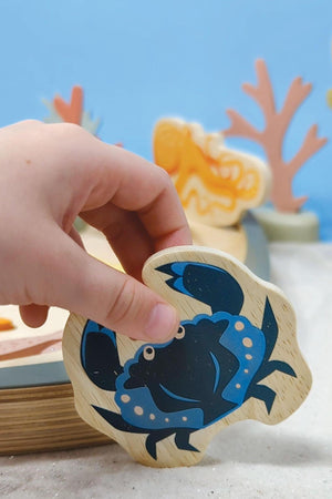 A childs hand holds a blue crab wooden toy, showcasing its vibrant colors against a playful underwater-themed background with coral and other sea creatures.