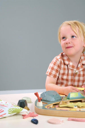 A young child with blonde hair sits at a table, engaging with a colorful wooden toy set featuring various shapes and textures, set against a neutral gray background.