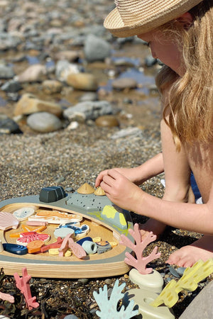 A child plays on a sandy beach with the Tender Leaf Wooden Toys My Little Rock Pool set, featuring colorful wooden sea creatures and rocks.