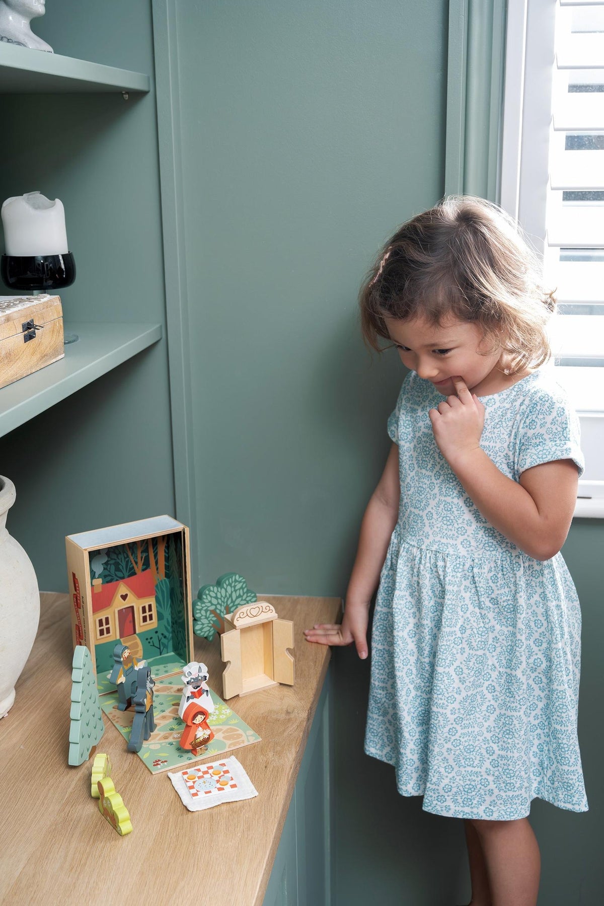 A young girl in a light blue dress examines a colorful wooden Little Red Riding Hood storytelling set, featuring characters and a woodland scene, displayed on a wooden shelf.