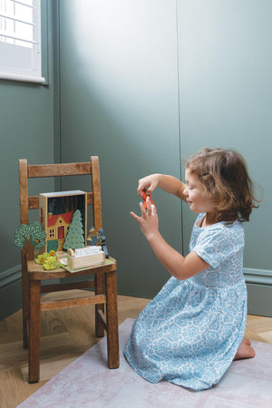 A young girl in a blue patterned dress plays with a wooden Little Red Riding Hood storytelling set on a small chair.