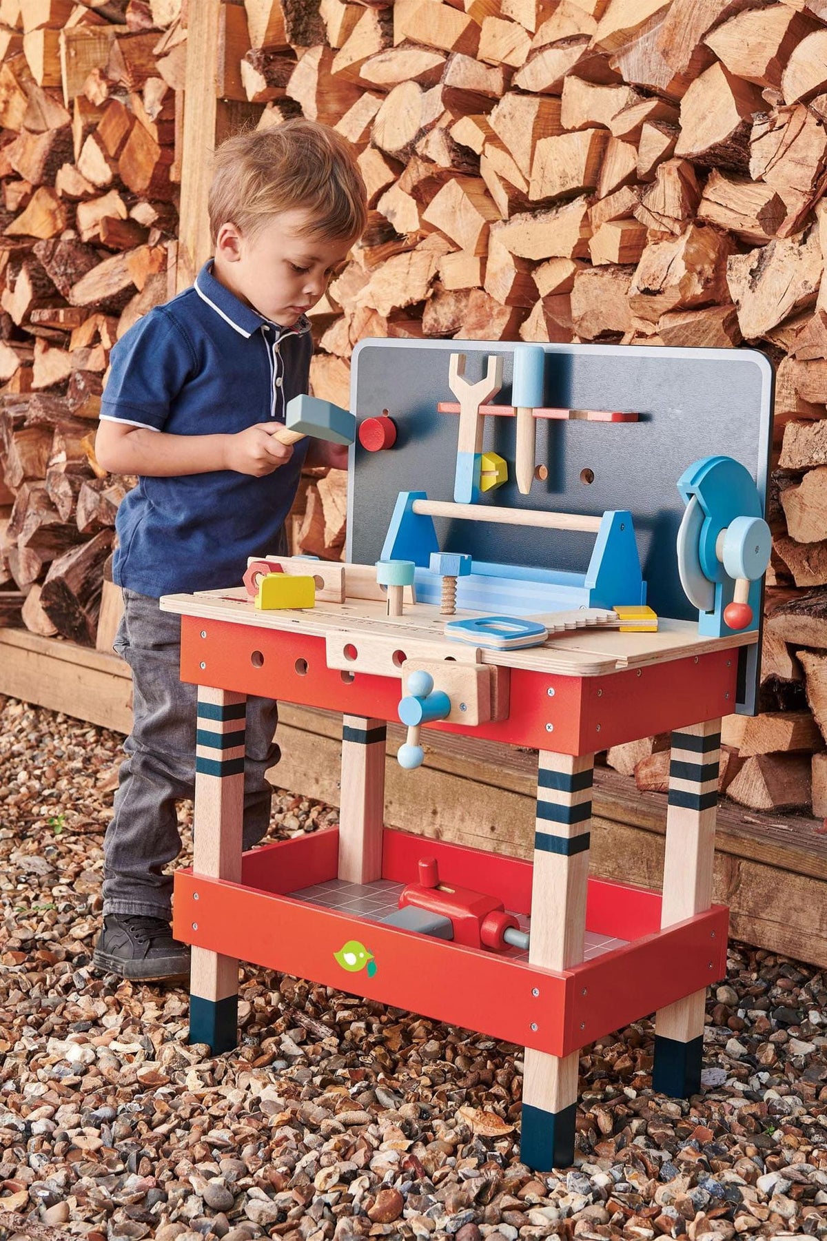 A young child plays at a colorful wooden tool bench outdoors, surrounded by stacked firewood, engaging with various toy tools.