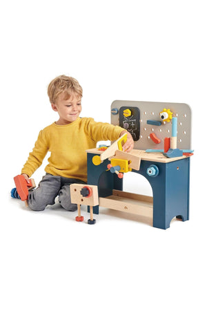 A young boy in a yellow sweater plays with a colorful wooden tool bench, featuring various tools and a blackboard.