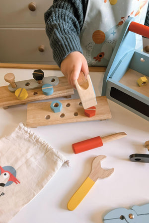 A child is holding a wooden piece above a colorful tool set, featuring various shapes and tools on a tabletop.