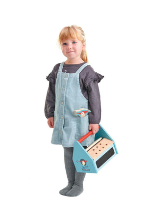 A young child in a blue corduroy dress holds a colorful wooden toolbox, featuring a playful woodpecker design.