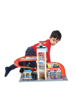 A child plays with a colorful Bluebird service station toy featuring ramps, a car wash, and a helipad.