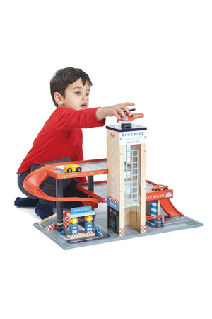 A child in a red shirt plays with a colorful wooden Bluebird service station toy featuring ramps and a car.