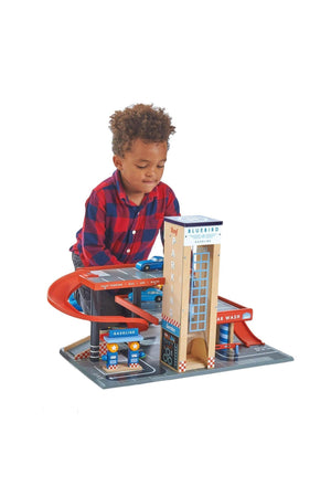 A child interacts with a colorful wooden toy service station featuring ramps, a car wash, and parking areas.
