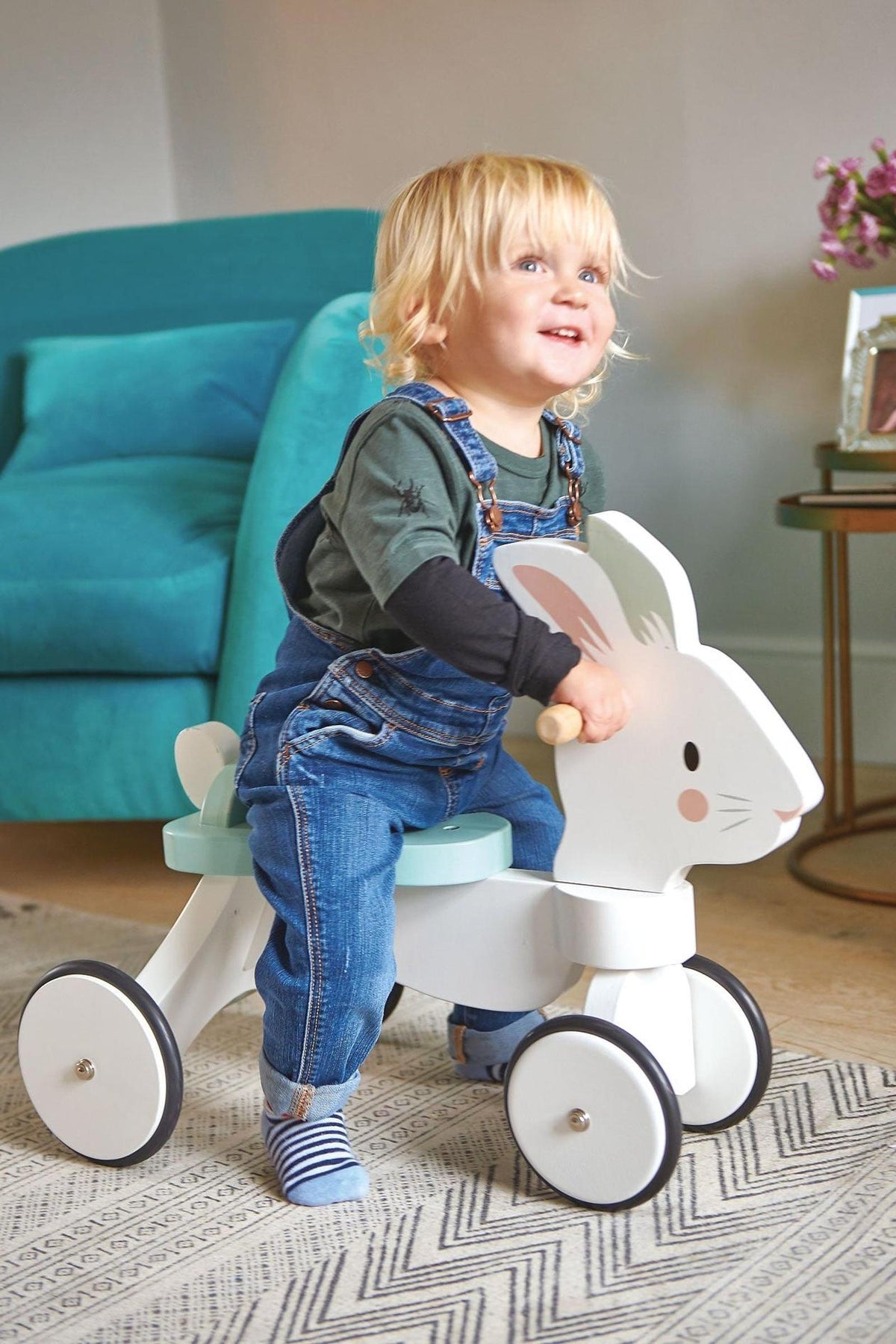A child with blonde hair smiles while riding a white wooden rabbit toy with rubber-trimmed wheels in a cozy living.