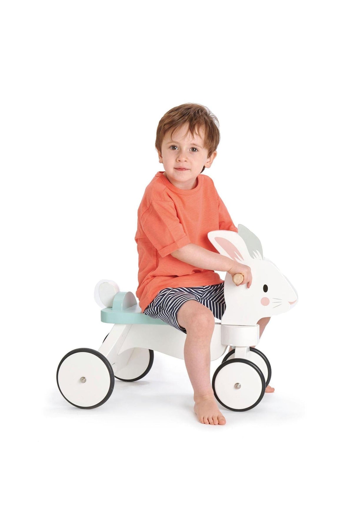 A young child sits on a white wooden ride-on rabbit toy with rubber wheels, wearing an orange shirt and striped.
