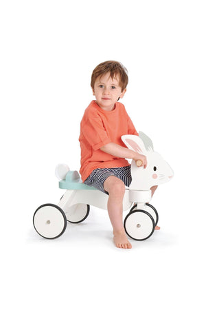 A young child sits on a white wooden ride-on rabbit toy with rubber wheels, wearing an orange shirt and striped.