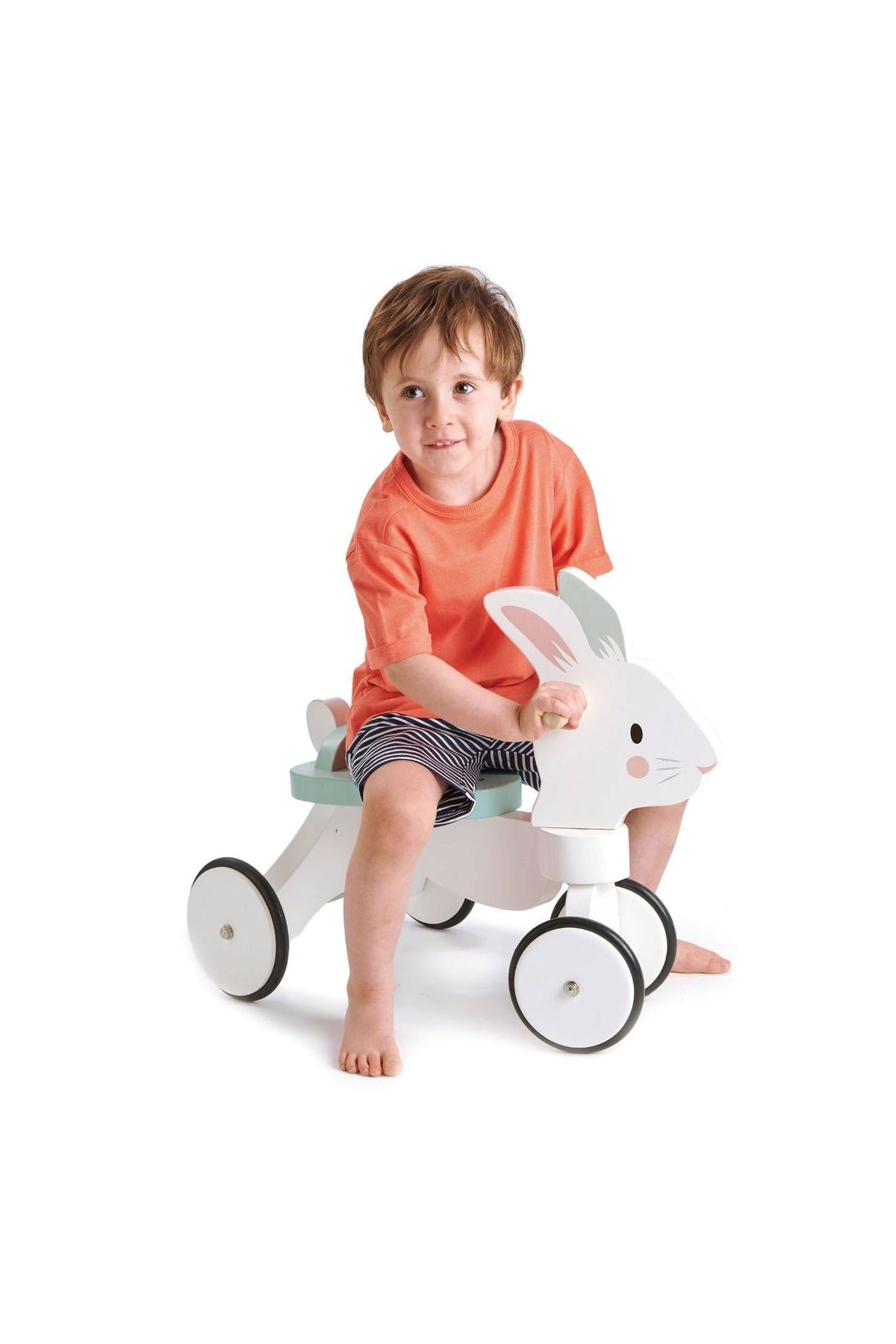 A young child sits on a white wooden ride-on bunny toy with non-slip wheels, wearing an orange shirt and striped.