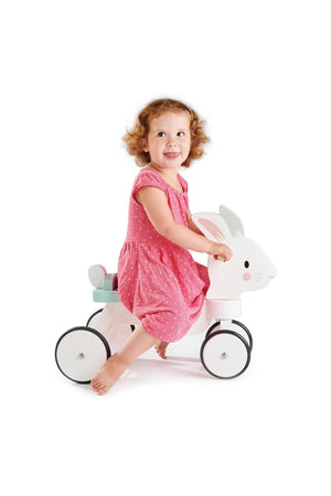 A young child in a pink dress rides a white wooden bunny toy with non-slip wheels, smiling happily.