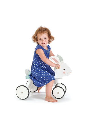 A young child smiles while sitting on a white wooden ride-on bunny toy, wearing a blue polka dot dress.