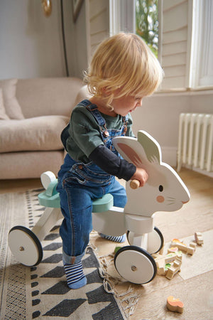 A child in denim overalls rides a white wooden bunny toy, surrounded by colorful wooden blocks on a patterned rug.