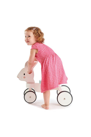 A toddler in a pink dress plays with a white wooden ride-on bunny toy, featuring black wheels and a friendly.