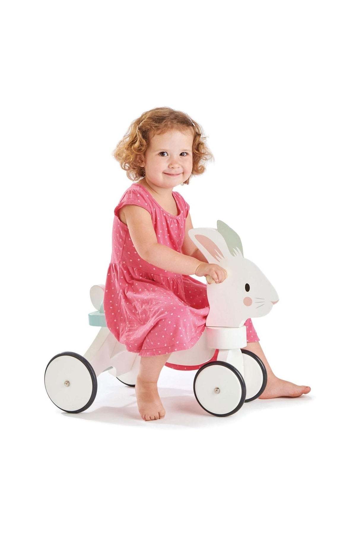 A child with curly hair smiles while riding a white wooden bunny toy with non-slip wheels, wearing a pink dress.