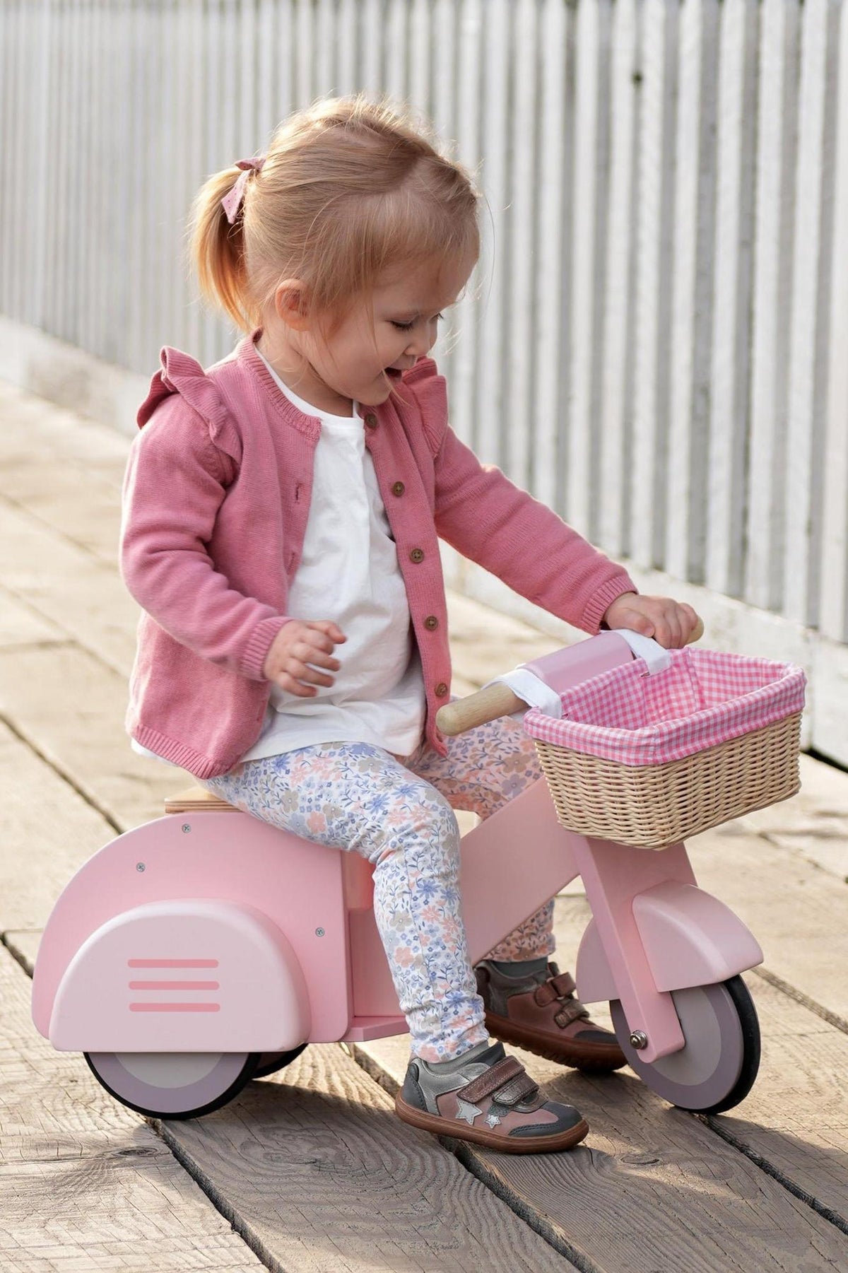 A young girl in a pink cardigan rides a wooden pink scooter with a wicker basket, smiling on a wooden.