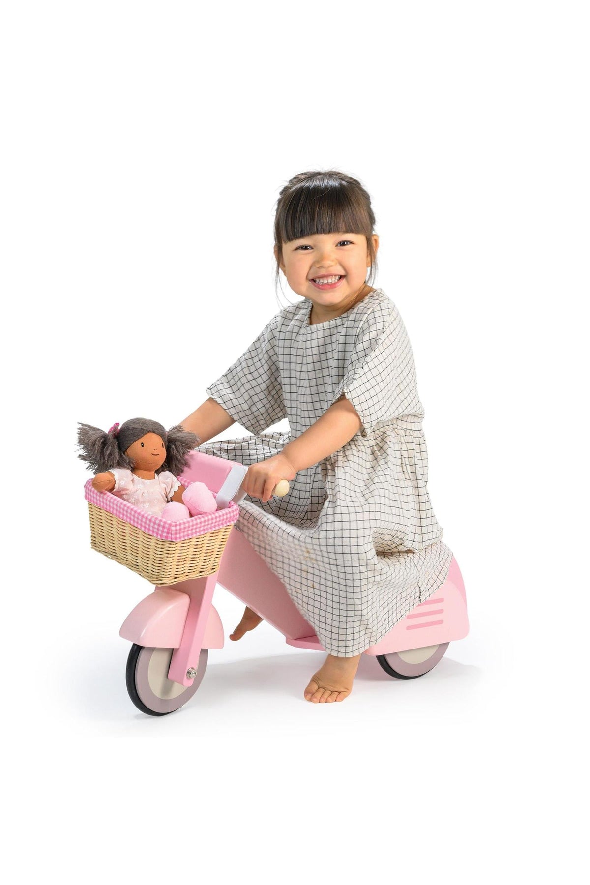 A smiling child in a checkered dress rides a pink wooden scooter with a wicker basket, carrying a doll.