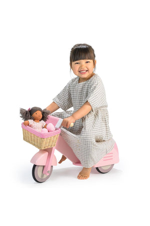 A smiling child in a checkered dress rides a pink wooden scooter with a wicker basket, carrying a doll.