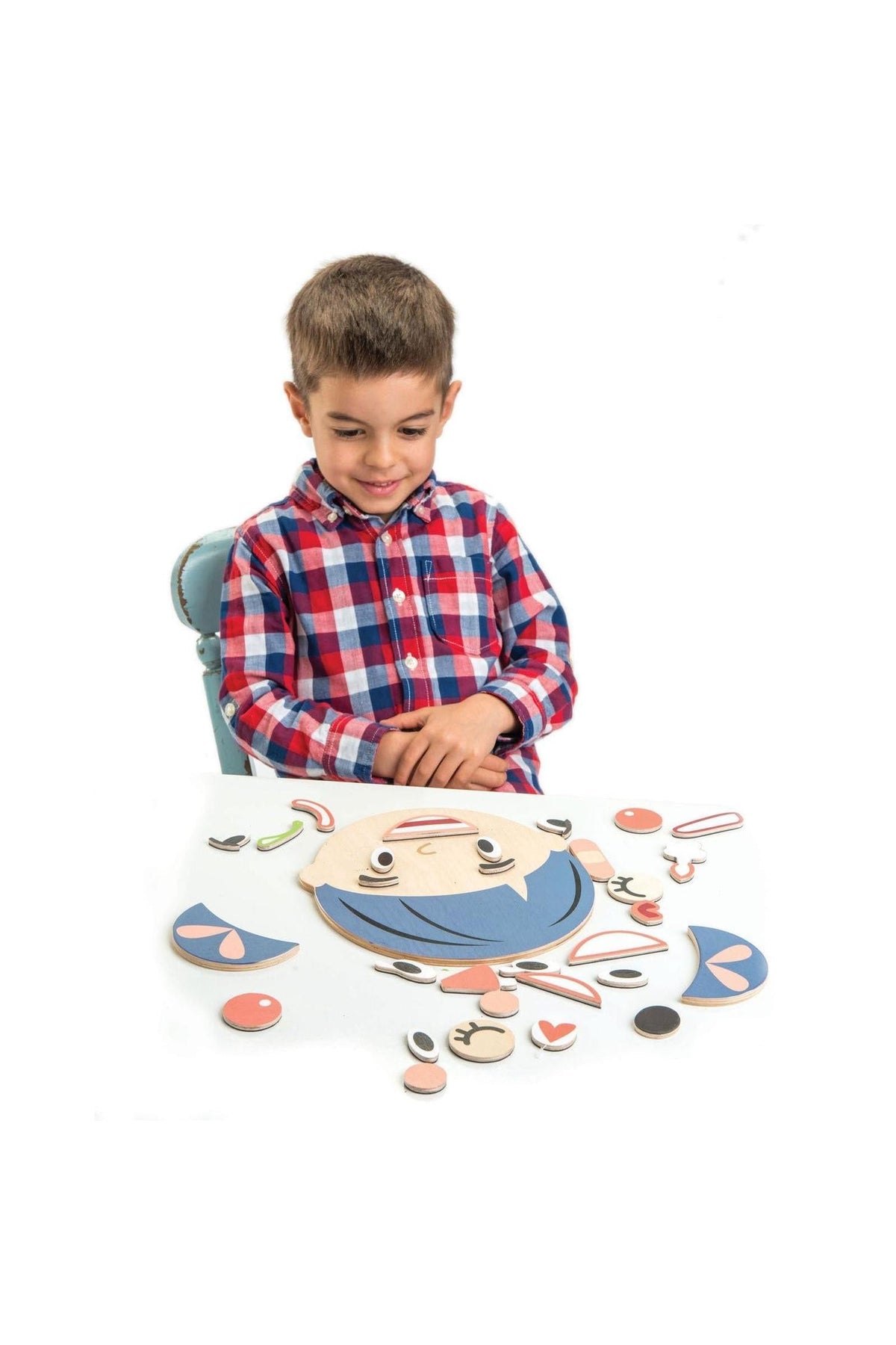 A young boy in a checkered shirt smiles while playing with wooden facial pieces on a table, creating various expressions.