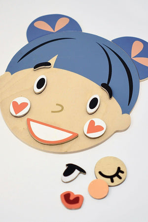 A wooden face-shaped base features blue hair buns and various magnetic facial pieces expressing emotions, promoting social development.