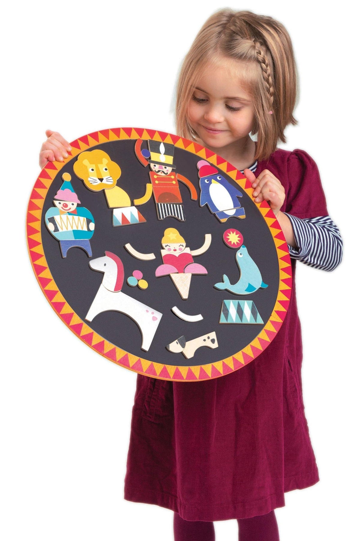 A young girl in a maroon dress holds a circular black board featuring colorful wooden circus characters, including a lion,.
