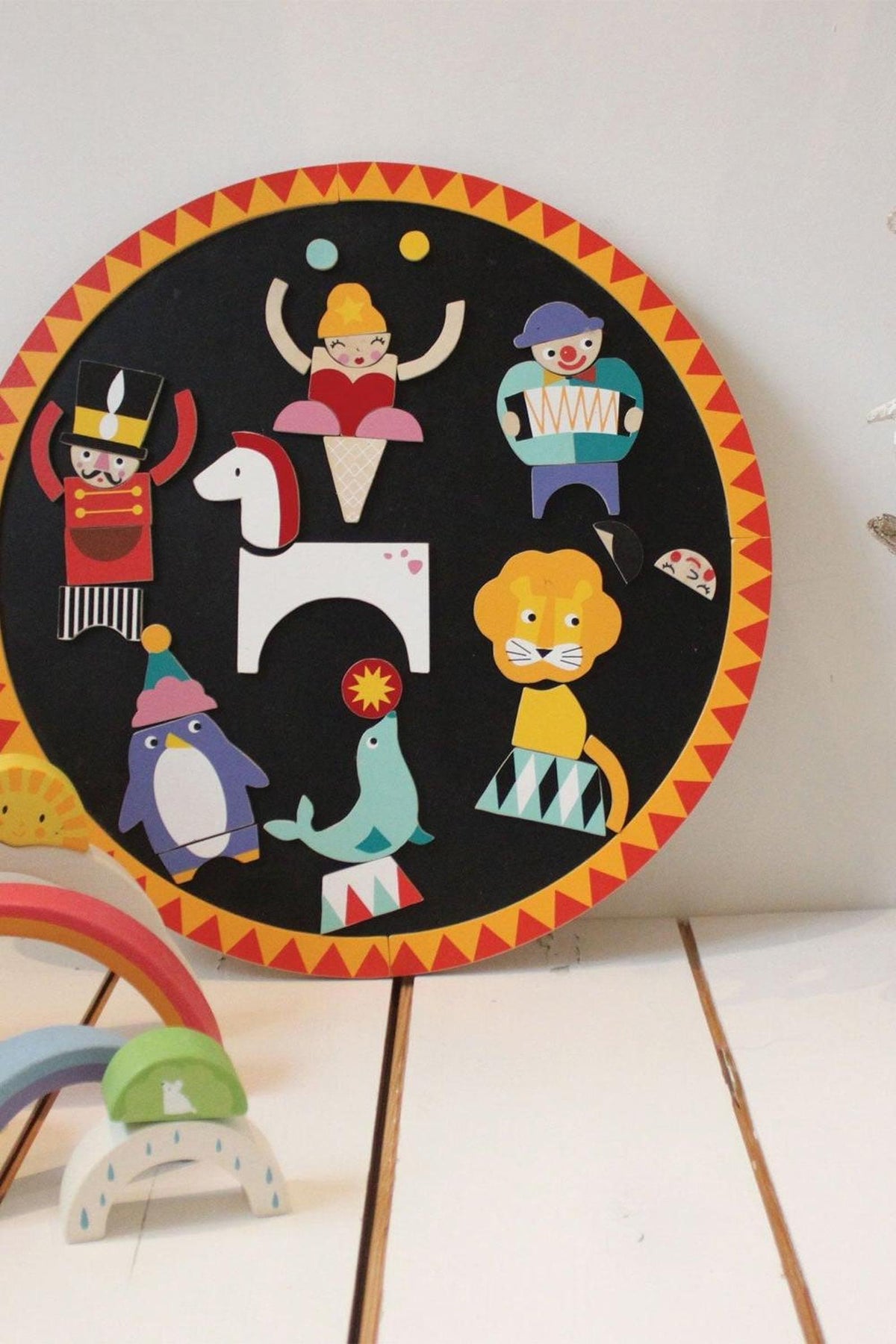 Colorful wooden magnetized circus characters are arranged on a circular black board, featuring a lion, penguin, and more.