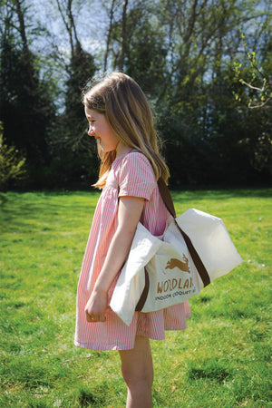 A young girl in a pink striped dress holds a canvas bag labeled Woodland Indoor Croquet while standing on grass.