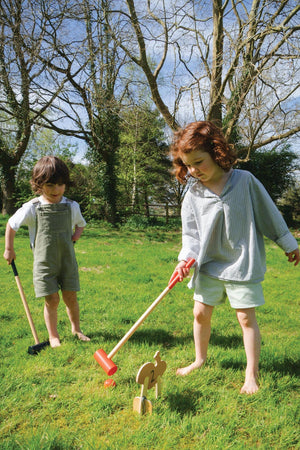 Two children play outdoors on grass, using wooden mallets in a croquet game with colorful, woodland-themed hoops.