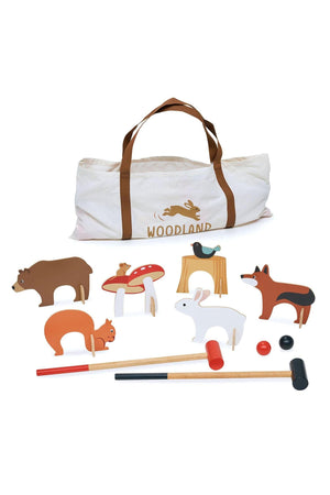 A printed canvas bag with brown straps contains colorful wooden croquet pieces, including woodland animals and two mallets.