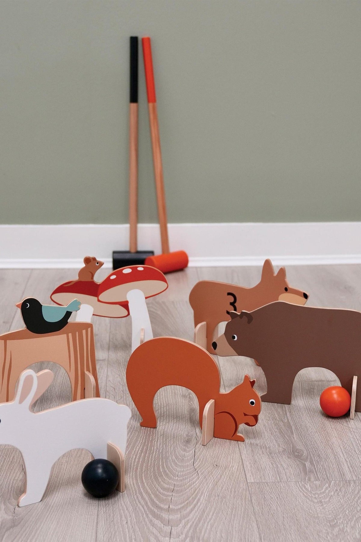 Colorful wooden croquet pieces featuring woodland animals and a toadstool, with two mallets in the background on a wooden floor.