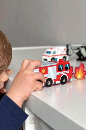 A child plays with a red wooden fire engine toy on a white surface, surrounded by emergency vehicle toys. The scene highlights engaging, sustainable play with quality materials.