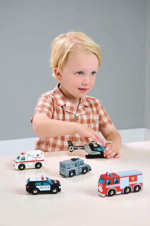 A young child plays with a set of colorful wooden emergency vehicles, including a police car, ambulance, fire engine, and S.W.A.T. truck, on a light-colored table.