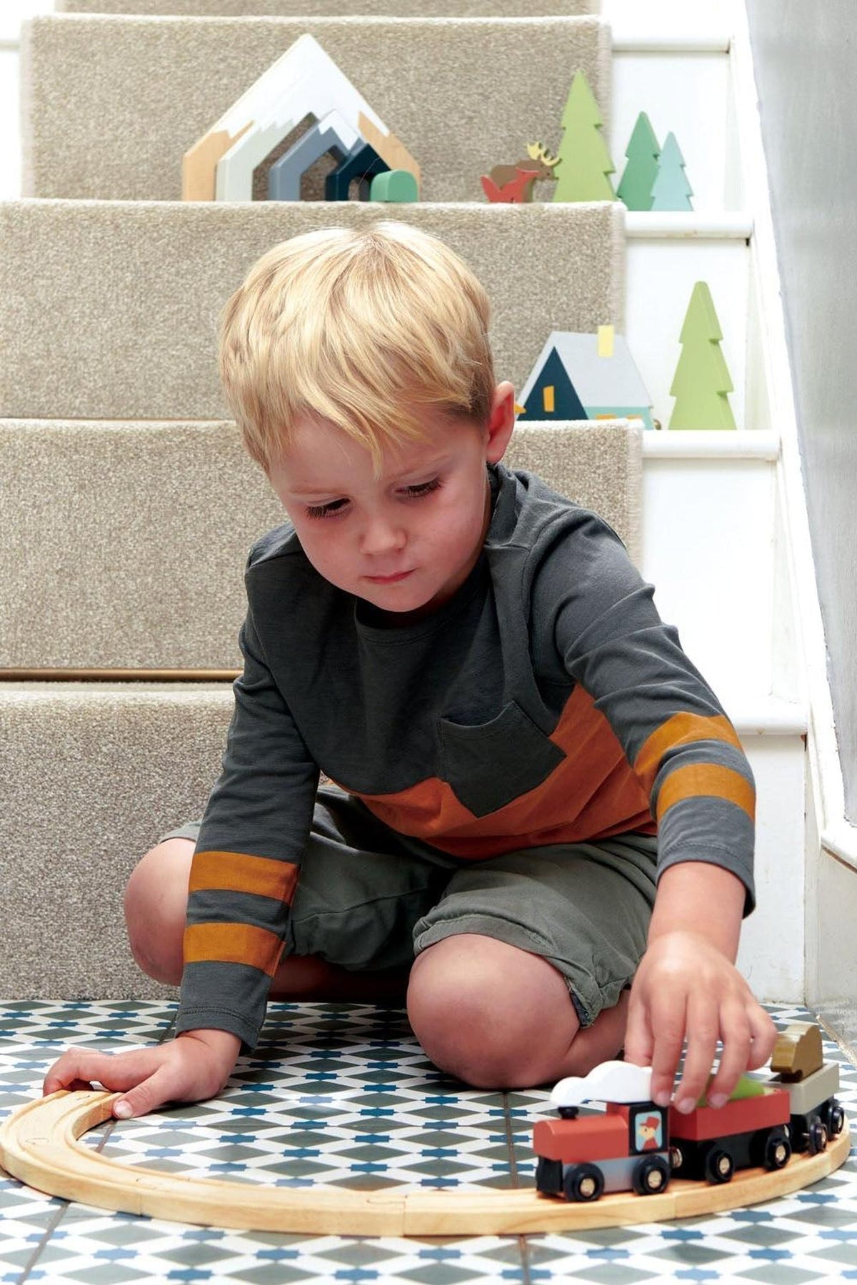 A young child plays on a patterned floor with a wooden train set, focusing intently on the trains carriages.