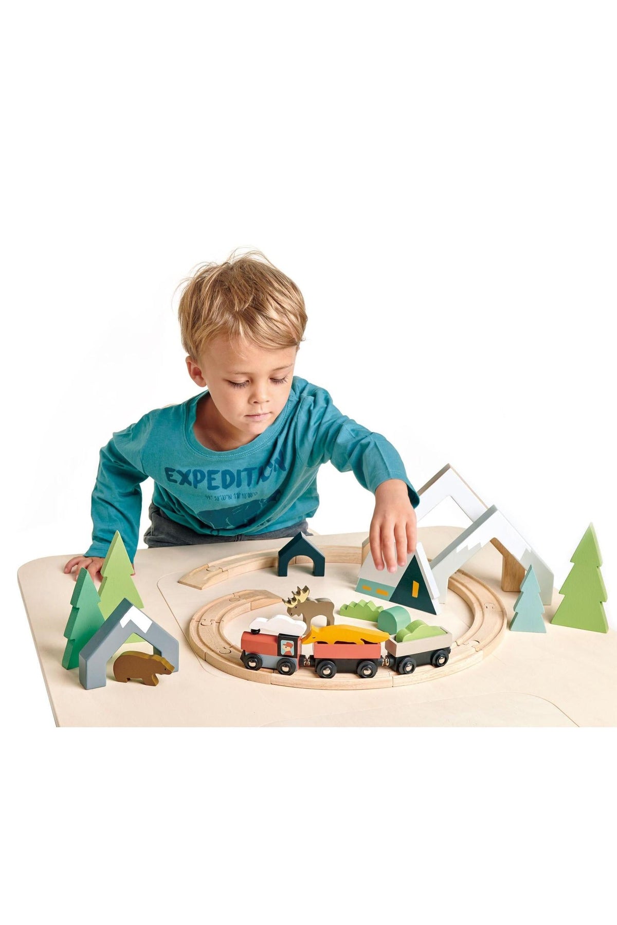 A young child plays with a wooden train set featuring colorful vehicles, trees, and a chalet on a light-colored table.