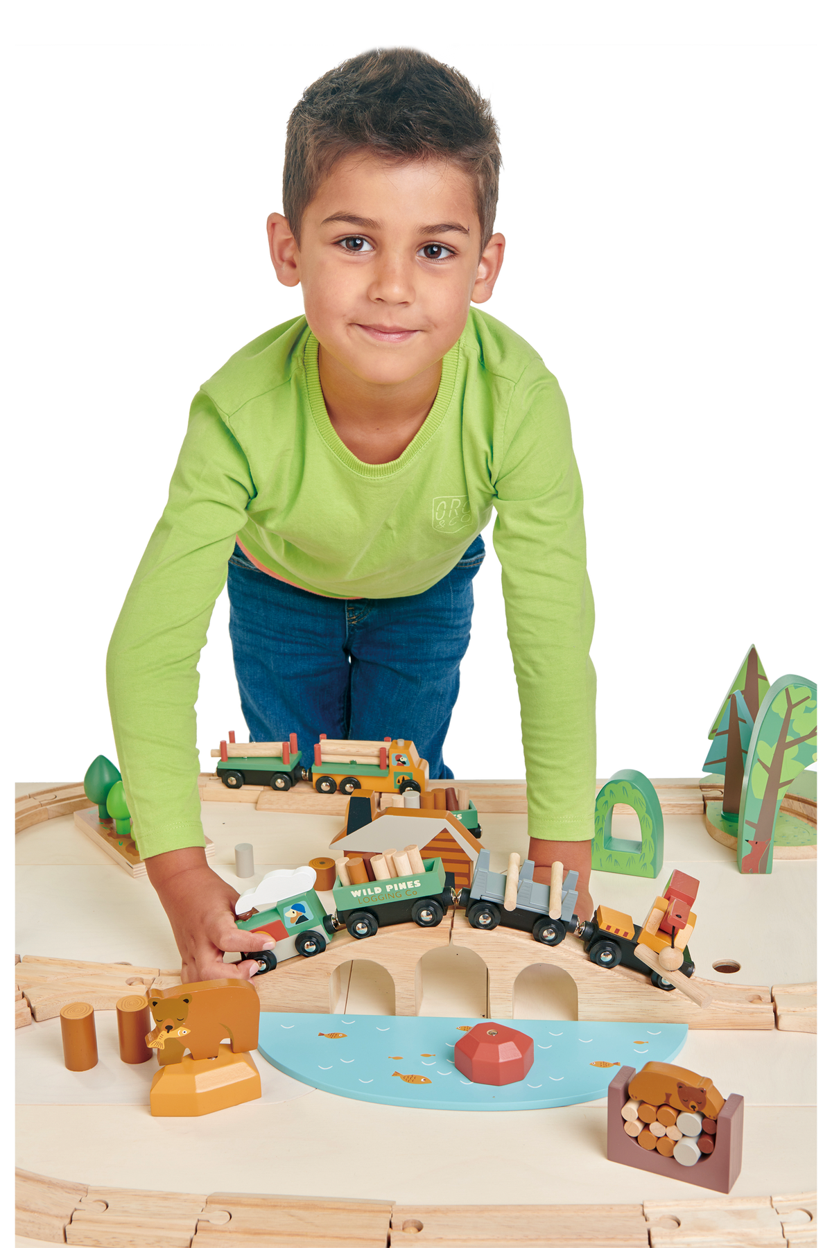 A young boy in a green shirt plays with a colorful wooden train set featuring various vehicles, trees, and logs on a light wooden table.
