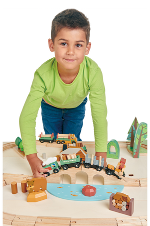 A young boy in a green shirt plays with a colorful wooden train set featuring various vehicles, trees, and logs on a light wooden table.