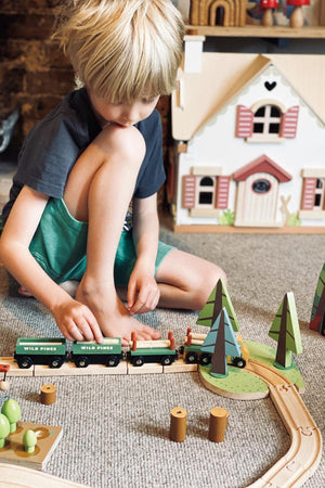 A young child plays with a Tender Leaf Wooden Toys Wild Pines Train Set on a carpeted floor, featuring colorful trees and train carriages, encouraging imaginative storytelling and creative play.