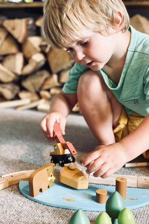A young child plays with a colorful wooden toy set, featuring an animal and a train on a blue base. The background showcases stacked logs, enhancing the natural, playful atmosphere.