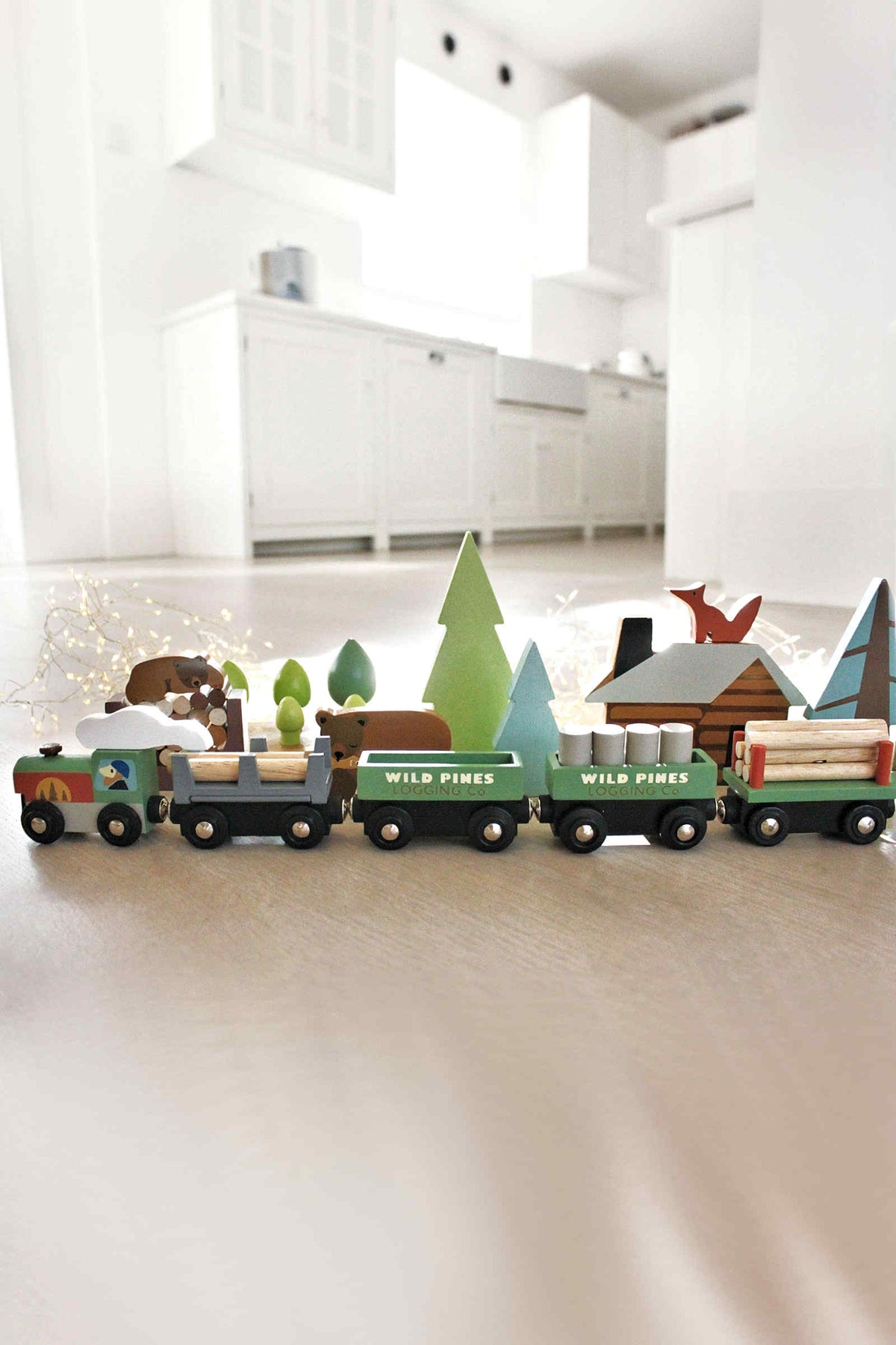 Colorful wooden train set with three carriages carrying logs, surrounded by pine trees and a cabin.