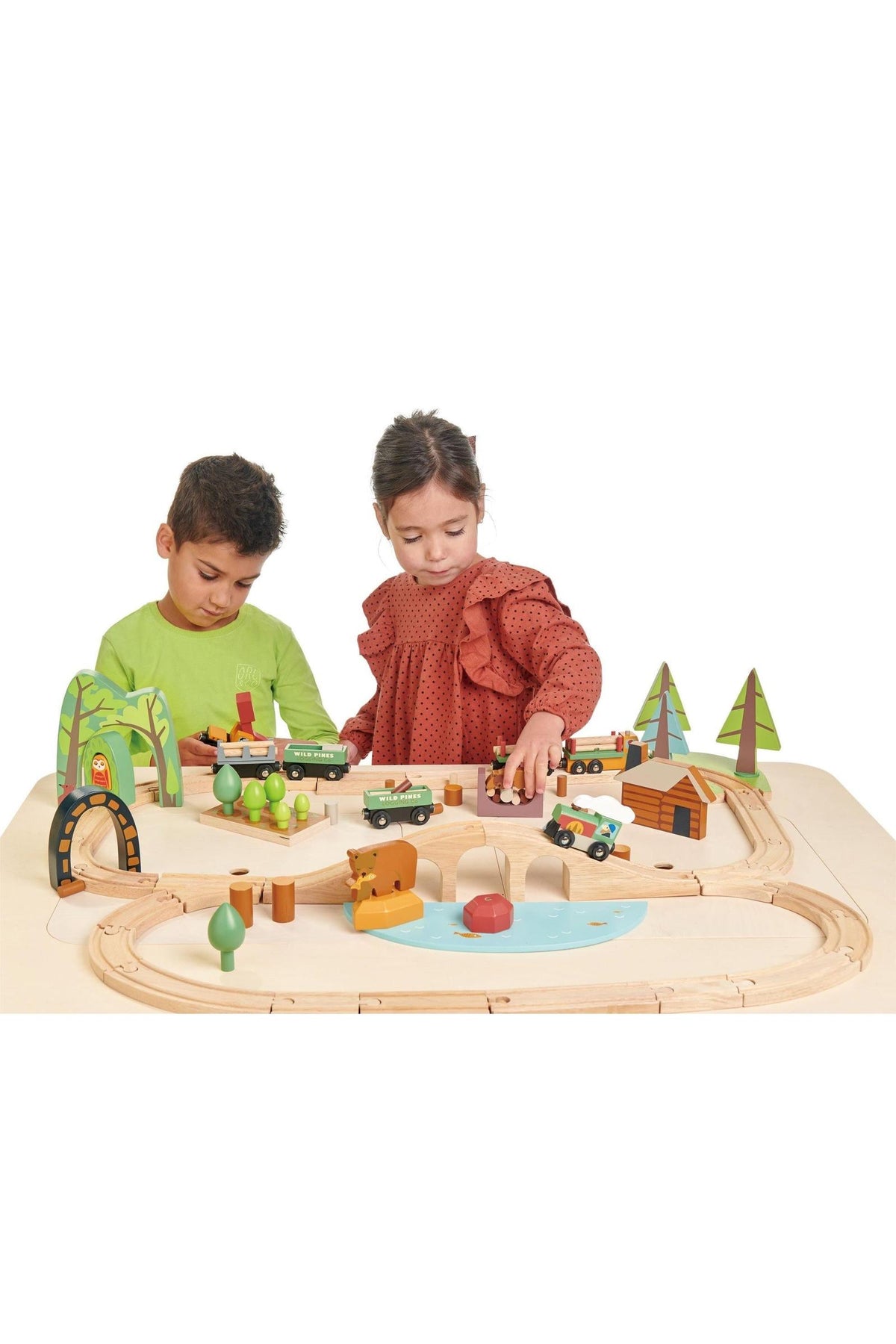 Two children play with a colorful wooden train set on a light wooden table. The set features trees, vehicles, and a variety of accessories, promoting imaginative play and storytelling.