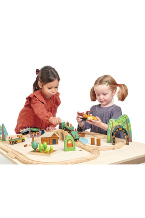 Two young children play with a colorful wooden train set, featuring trees and a bridge, on a light wooden table. The scene encourages imaginative storytelling and interactive play.